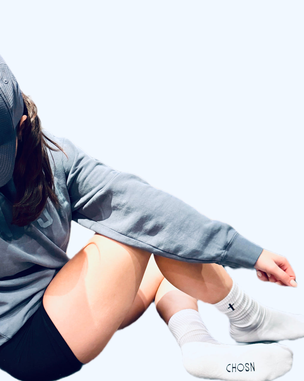 Person wearing a gray hoodie and white socks with a cross and the letters CHOSN  on the bottom of sock sitting on a light surface.