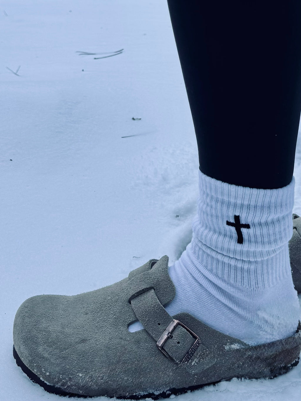 Gray clog shoe worn with a white sock featuring a black cross design on a snowy ground.