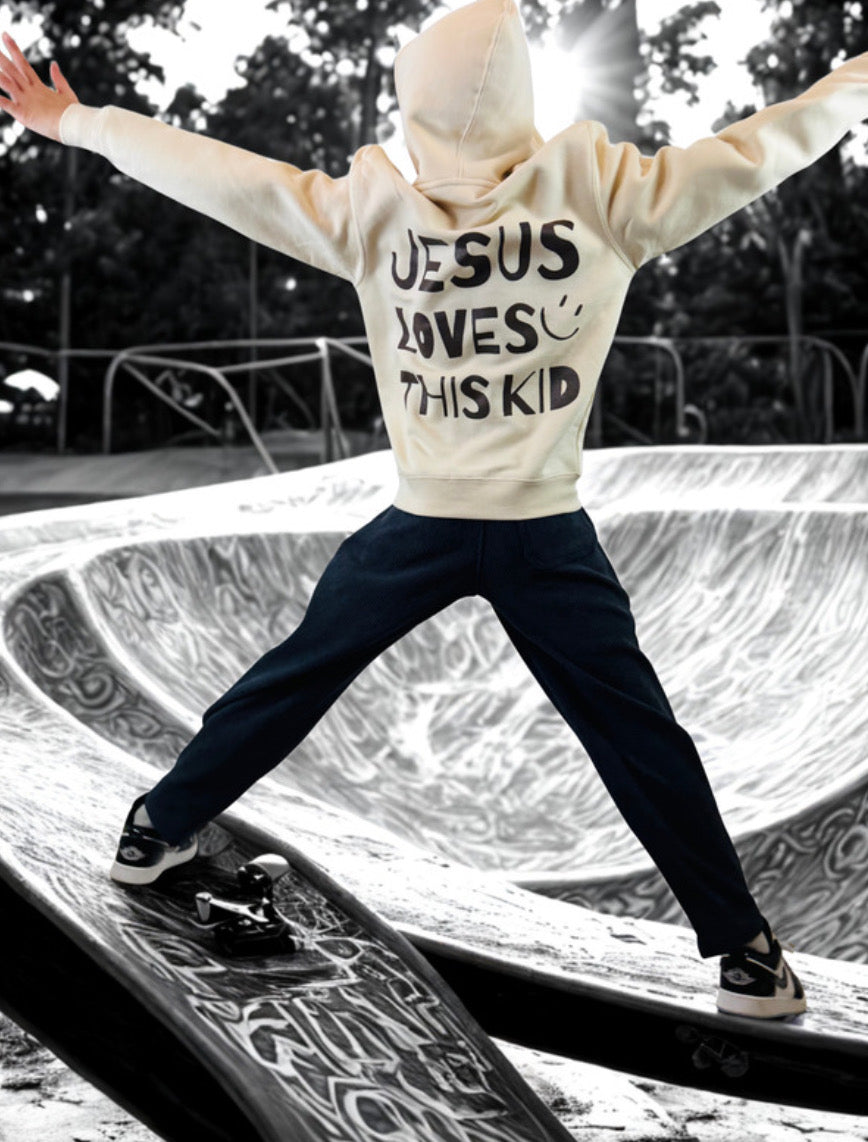 Youth wearing a cream hoodie with 'Jesus Loves This Kid' in Chocolate Brown text at a skate park.