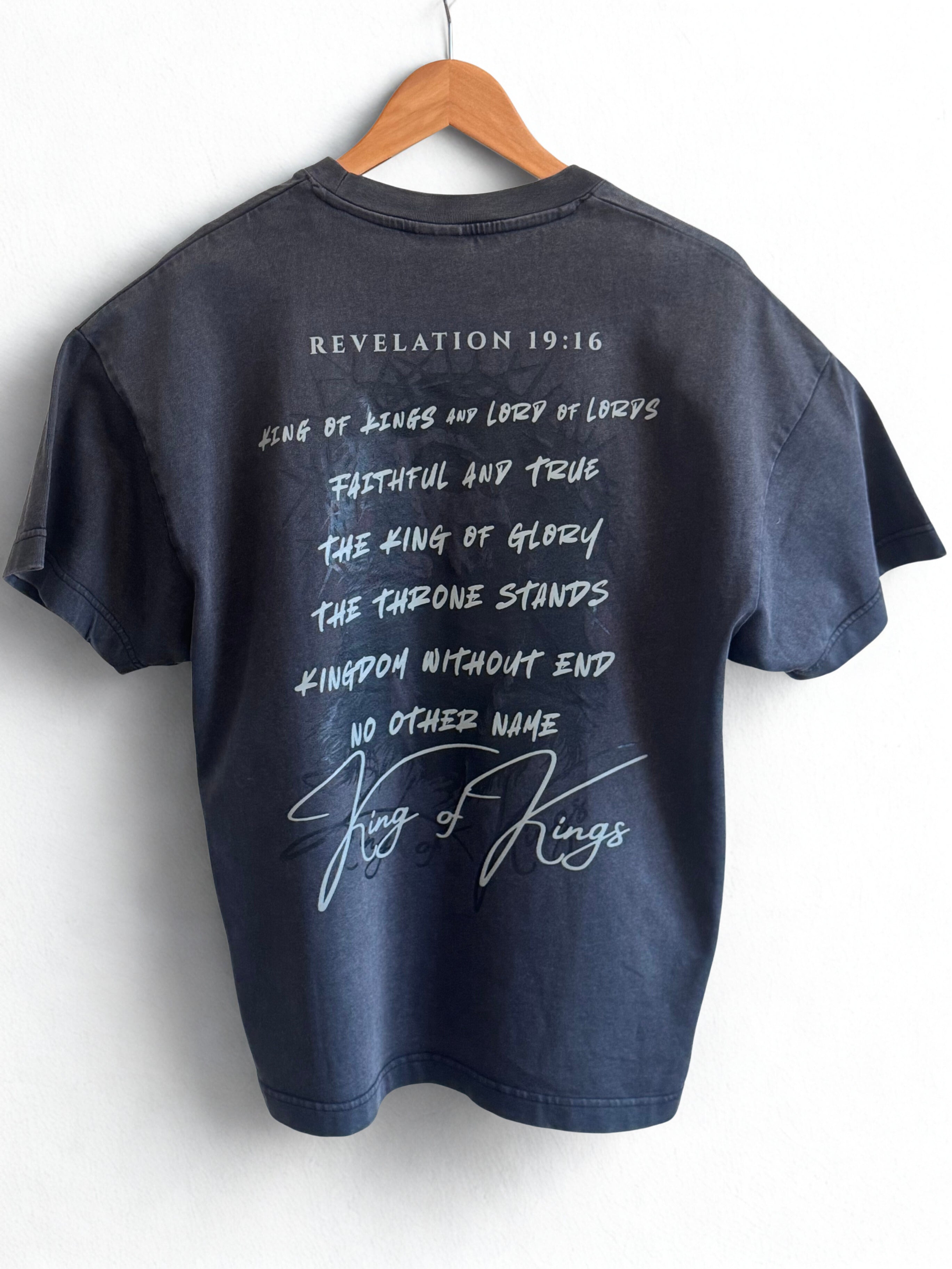 Back of black vintage t-shirt with "Revelation 19:16 and religious text reading "King of Kings and Lord of Lords Faithful and True The King of Glory The Throne Stands Kingdom without End No other Name King of Kings"printed in light grey lettering over top a faded black image of Jesus' face. 
