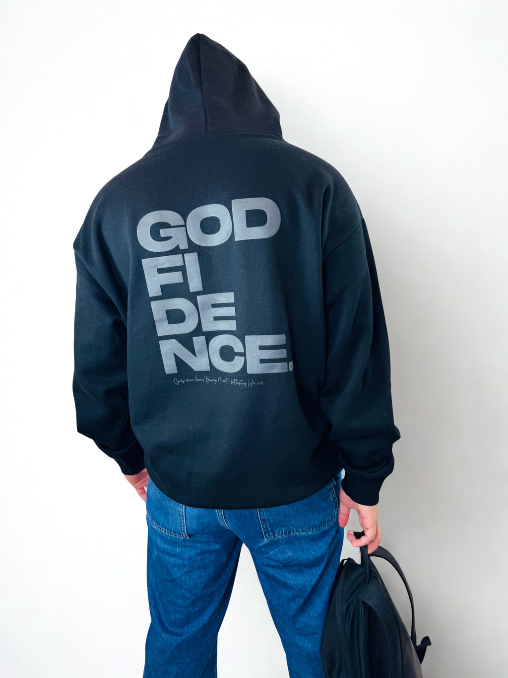 Person wearing a black hoodie with 'GOD FIDENCE' text on the back, holding a black bag.