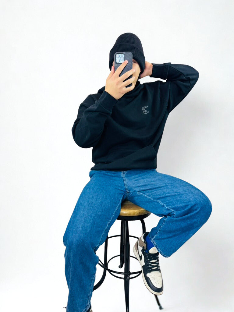 Person wearing a black hoodie and blue jeans sitting on a stool against a white background